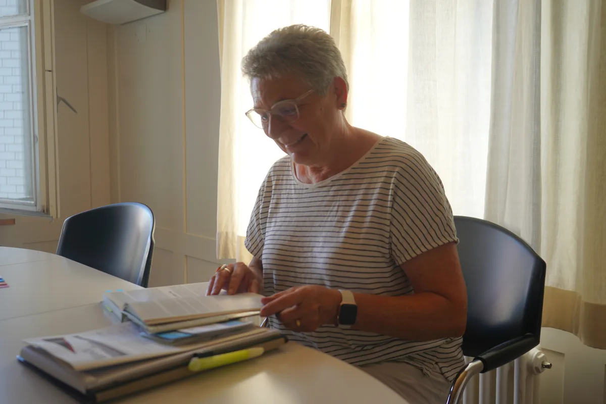 Eine Frau sitzt in einem Zimmer vor einem Tisch und blättert in einem Buch.